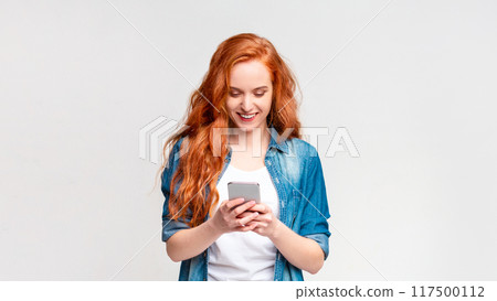 A woman with vibrant red hair is intensely focused on her cell phone screen. She appears engrossed in whatever she is viewing, holding the device with both hands A woman with vibrant red hair is intensely focused on her cell phone screen. She appears engrossed in whatever she is viewing, holding the device with both hands 117500112