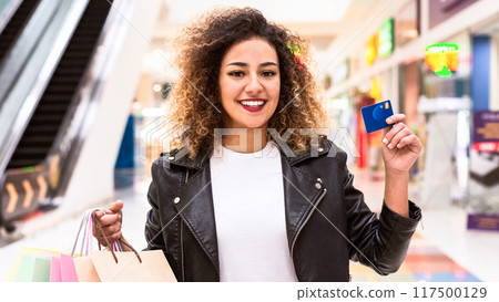Pretty African American woman stands in a bustling shopping mall, holding a credit card in her hand. She appears to be making a purchase or preparing to do so. Pretty African American woman stands in a bustling shopping mall, holding a credit card in her hand. She appears to be making a purchase or preparing to do so. 117500129