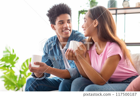 African American teen guy and girl are sitting on a couch in a home, each holding a white coffee mug, looking at each other and smiling, enjoying a moment of intimacy and connection over coffee. 117500187
