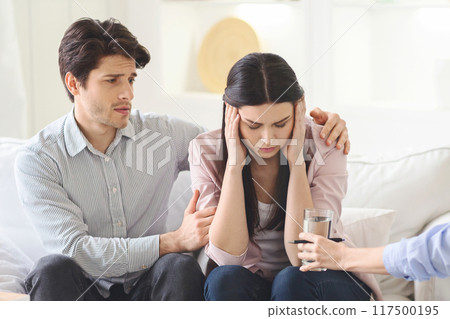 A worried man places a comforting arm around a distressed woman sitting on a sofa. They are indoors during the daytime, and another person offers a glass of water, counselor office 117500195
