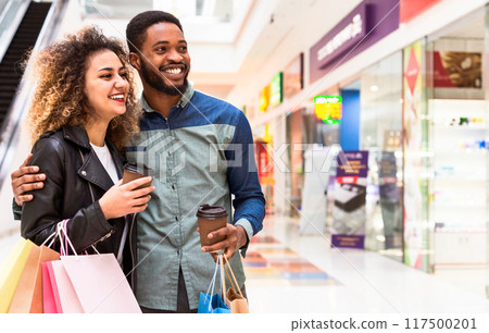 A cheerful black man and woman walk through a mall carrying shopping bags and coffee cups, sharing a conversation and a laugh together, showcasing a moment of casual leisure and retail therapy. 117500201