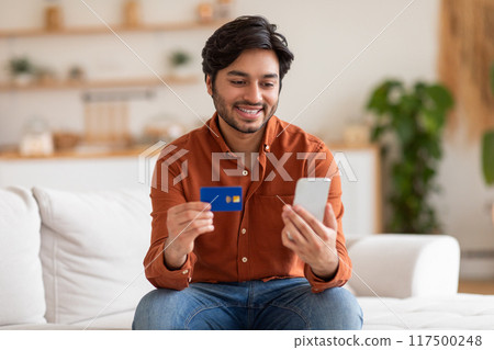 A young man is sitting on a white couch, holding a credit card and a smartphone, smiling at the screen. He is making an online purchase, likely shopping online or paying bills. 117500248