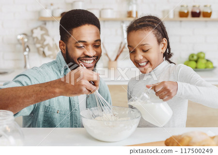 Cute African American Girl Pouring Milk Into Bowl At Kitchen, dad mixing the dough Cute African American Girl Pouring Milk Into Bowl At Kitchen, dad mixing the dough 117500249