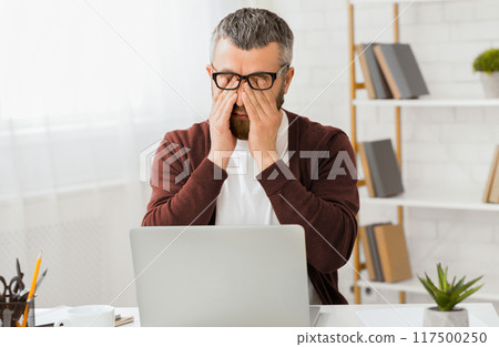 A middle aged man wearing glasses and a sweater sits at a desk in his home office. He is rubbing his eyes in frustration with an open laptop in front of him. Books and office supplies are arranged 117500250