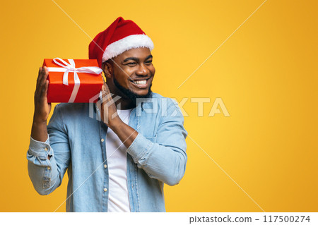Guess what's inside. Happy black man in Santa hat shaking Christmas present, listening what rattles in gift box, yellow background Guess what's inside. Happy black man in Santa hat shaking Christmas present, listening what rattles in gift box, yellow background 117500274
