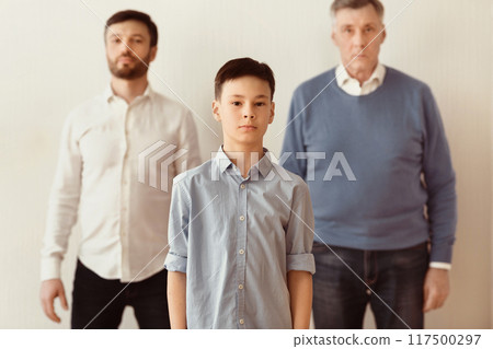 Male Generations Concept. Serious Boy Standing Between Dad And Grandpa Against White Wall Indoor. Selective Focus Male Generations Concept. Serious Boy Standing Between Dad And Grandpa Against White Wall Indoor. Selective Focus 117500297