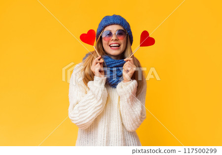 Smiling pretty winter girl in knitted set and sunglasses with red heart cards over pink studio background, be my valentine Smiling pretty winter girl in knitted set and sunglasses with red heart cards over pink studio background, be my valentine 117500299