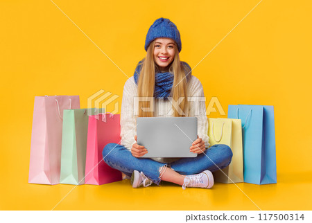 Pretty winter girl in warm hat and scarf with laptop sitting on floor among shopping bags, pink background, copy space 117500314