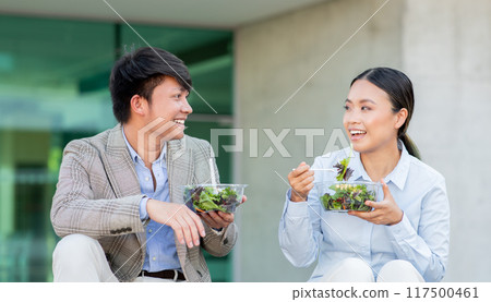 Two young Asian adults man and woman are seated on a stone surface, sharing a light and healthy lunch consisting of fresh salads. They are smiling and engaged in a cheerful conversation Two young Asian adults man and woman are seated on a stone surface, sharing a light and healthy lunch consisting of fresh salads. They are smiling and engaged in a cheerful conversation 117500461