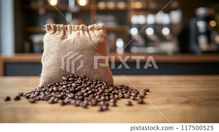 A burlap bag filled with coffee beans is spilled on a rustic wooden table inside a warm cafe, showcasing the rich texture and color of the beans. 117500525