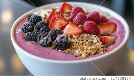 A vibrant smoothie bowl features a blend of berries, topped with granola, strawberries, and raspberries, served in a cozy cafe. 117500534