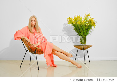 Elegant woman in a flowing coral dress lounges next to vibrant yellow flowers 117500796