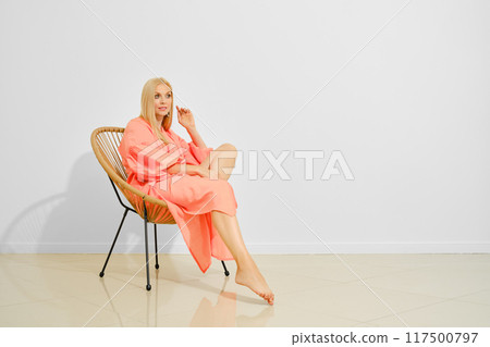 Cute woman in coral maxi dress sits in wicker chair in bright room 117500797