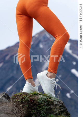 Athletic female legs wearing orange body hugging workout leggings and white sneakers. Focuses on lower part of female sportsperson body during outdoor fitness training in mountains in background 117500981