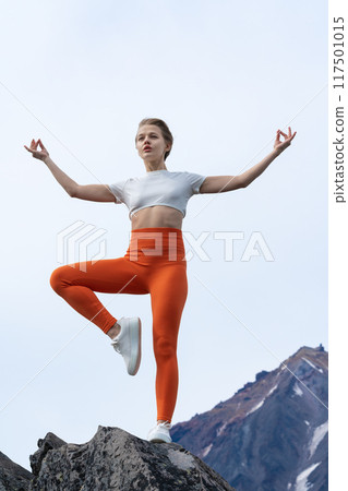 Flexible female yoga instructor balancing her body on one leg while doing yoga, standing on top of rocky mountain with volcano in background. Slim woman wears orange yoga leggings and sneakers 117501015