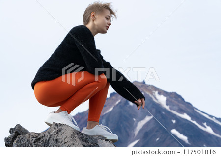 Woman tourist sits squatting on rocky outcrop in mountains during summer travel through mountainous area with volcano cone in background. Lonely woman enjoying moment of tranquility, looking at view 117501016