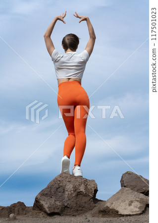 Rear view of woman practicing yoga, standing on rocky mountaintop on background of sky with clouds. Back view of body of slender female athlete. Yoga instructor in T-shirt, yoga leggings and sneakers Rear view of woman practicing yoga, standing on rocky mountaintop on background of sky with clouds. Back view of body of slender female athlete. Yoga instructor in T-shirt, yoga leggings and sneakers 117501023