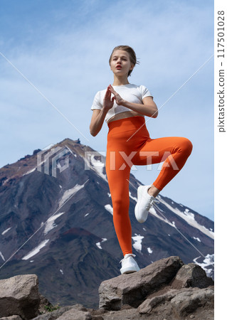 Woman practices yoga standing on one leg against backdrop of volcano. Shapely woman balance body vital Zen meditation for workout. Yoga trainer wearing t-shirt, orange yoga leggings, sneakers 117501028