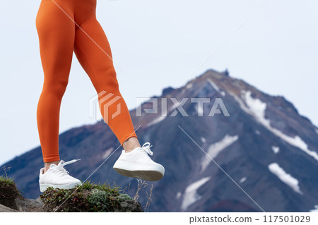 Unrecognizable woman doing fitness workout in mountains with volcano in background. Close-up of legs in orange figure hugging sports leggings and white sneakers. Low section of female muscular body 117501029