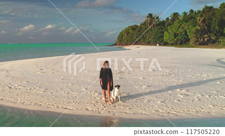 Young woman is enjoying a relaxing walk with her dog on a pristine white sand beach, with lush tropical vegetation and turquoise waters in the background 117505220