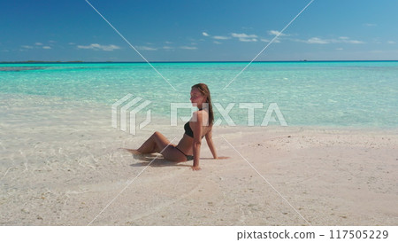 Young woman enjoys the sun and crystal clear water while sitting on a sandbank. The turquoise ocean stretches as far as the eye can see 117505229