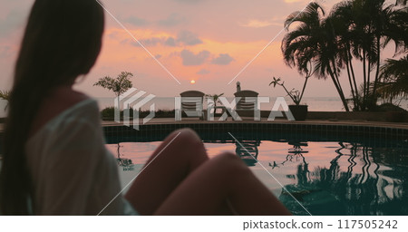 A woman is seated at the edge of a pool in Thailand, contemplating the water below her. Bright sunset in background 117505242