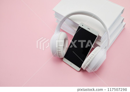 Audio books concept. Headphones and smartphone on a stack of white books over a pink background. Audio books concept. Headphones and smartphone on a stack of white books over a pink background. 117507298