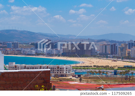 Panoramic landscape of the city with a sandy beach with vacationers on the shore with urban development, mountains and clouds with blue sky 117509265