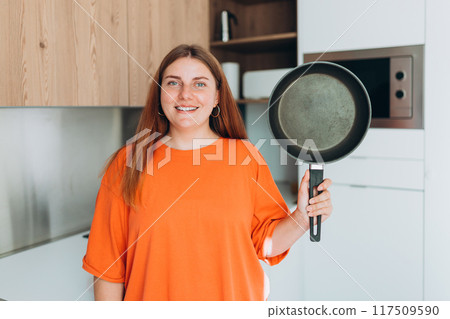 Young woman housewife holding empty pan in the kitchen at home. Domestic life and diet concept. Food banner. Time to eat Young woman housewife holding empty pan in the kitchen at home. Domestic life and diet concept. Food banner. Time to eat 117509590