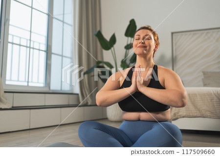 Yoga mindfulness meditation. Woman practicing yoga in living room at home. Plus size female sitting in lotus pose meditating smiling relaxing indoor. Girl doing breathing practice. Yoga at home Yoga mindfulness meditation. Woman practicing yoga in living room at home. Plus size female sitting in lotus pose meditating smiling relaxing indoor. Girl doing breathing practice. Yoga at home 117509660