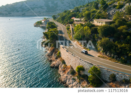 Car with paddleboard on the roof drives along a narrow coastal road under a clear blue sky. 117513016