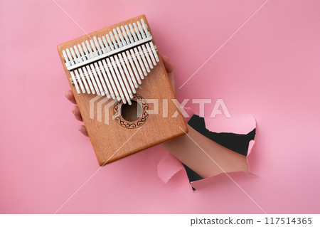 Female hand holding a kalimba through a hole in pink paper Female hand holding a kalimba through a hole in pink paper 117514365