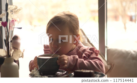 A girl drinks hot cocoa from a green cup against the backdrop of a cozy cafe with large panoramic windows. 117514441