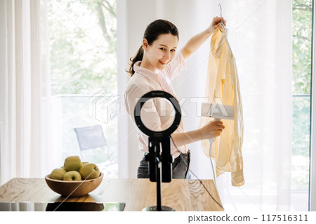 Young woman using a handheld steamer to smooth out a shirt while recording a vlogging tutorial at home, highlighting DIY fashion tips and content creation. 117516311