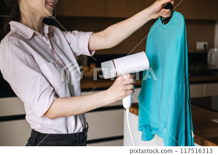 Woman steaming a blue shirt with handheld steamer at home. Garment care, laundry, wrinkle removal, home chores, clothing maintenance. Woman steaming a blue shirt with handheld steamer at home. Garment care, laundry, wrinkle removal, home chores, clothing maintenance. 117516313