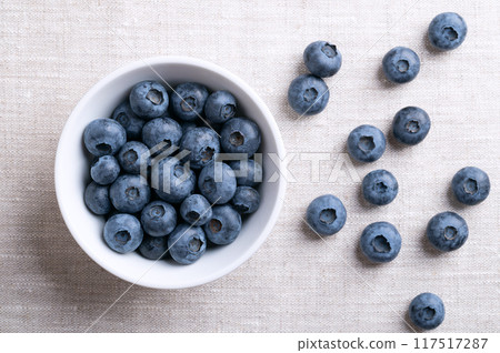 Blueberries in a white bowl on linen fabric. Dark blue colored, ripe, raw berries, fruits of Vaccinium corymbosum, cultivated blueberries of the northern highbush blueberry. From above, food photo. 117517287