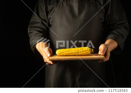 The waiter serves a head of boiled corn on a wooden cutting board in his hands. Tasty snack concept on black background with space for advertising. Corn diet The waiter serves a head of boiled corn on a wooden cutting board in his hands. Tasty snack concept on black background with space for advertising. Corn diet 117518565