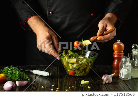 Chef preparing vegetable salad in tavern kitchen. Concept of preparing vegetable menu for lunch. Spoon in cook hand with fresh chopped vegetables Chef preparing vegetable salad in tavern kitchen. Concept of preparing vegetable menu for lunch. Spoon in cook hand with fresh chopped vegetables 117518566