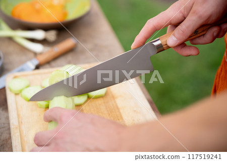 Close up of man holding sharp knife, chopping cucumber. Preparing vegetables for an outdoor barbecue. Close up of man holding sharp knife, chopping cucumber. Preparing vegetables for an outdoor barbecue. 117519241