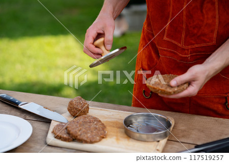 Man in apron preparing meat for hamburgers, standing outdoors. Using burger patty press to shape groud meat. Man in apron preparing meat for hamburgers, standing outdoors. Using burger patty press to shape groud meat. 117519257