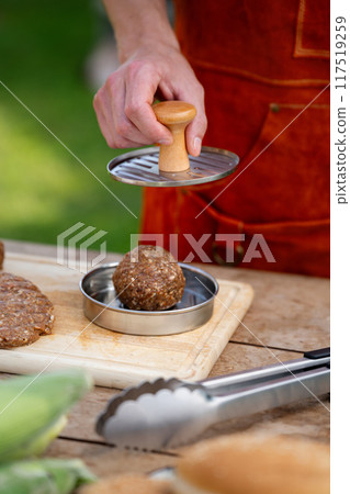 Man in apron preparing meat for hamburgers, standing outdoors. Using burger patty press to shape groud meat. 117519259