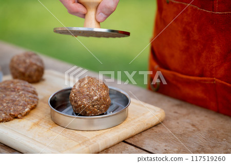 Man in apron preparing meat for hamburgers, standing outdoors. Using burger patty press to shape groud meat. 117519260