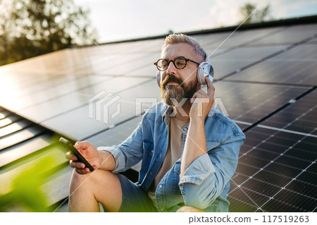 Man sitting on roof full of solar panels, headphones on head and closed eyes. Man sitting on roof full of solar panels, headphones on head and closed eyes. 117519263