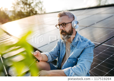 Man sitting on roof full of solar panels, headphones on head, holding smartphone. 117519264