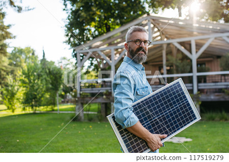 Man carrying solar panel in arms, walking across garden. Sustainable lifestyle and green household. Man carrying solar panel in arms, walking across garden. Sustainable lifestyle and green household. 117519279