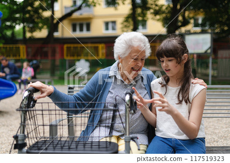 Grandma with walker picking up young girl from school at afternoon, spending time with senior grandmother in city park, sitting on bench in children playground. 117519323