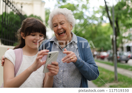 Grandma picking up young girl from school at afternoon. Granddaughter showing something on smartphone to senior grandmother. 117519330