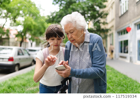 Grandma picking up young girl from school at afternoon. Granddaughter showing something on smartphone to senior grandmother, teaching her how to work with technology. 117519334