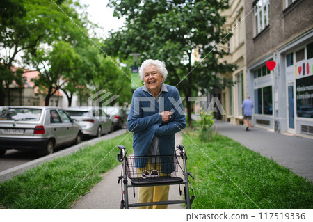 Beautiful elderly woman walking on city street with rollator, going shopping to the store. Feeling cold. 117519336