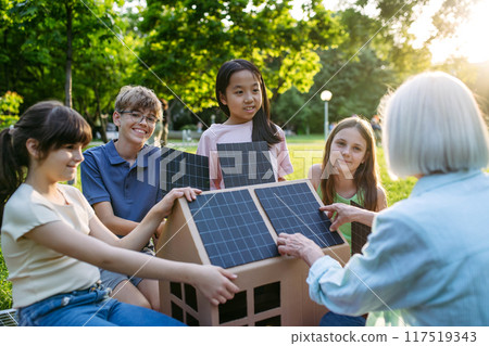 Children learning about renewable energy and solar panels during sustainable education class outdoors, using cardboard model of house wit solar panel on roof. 117519343